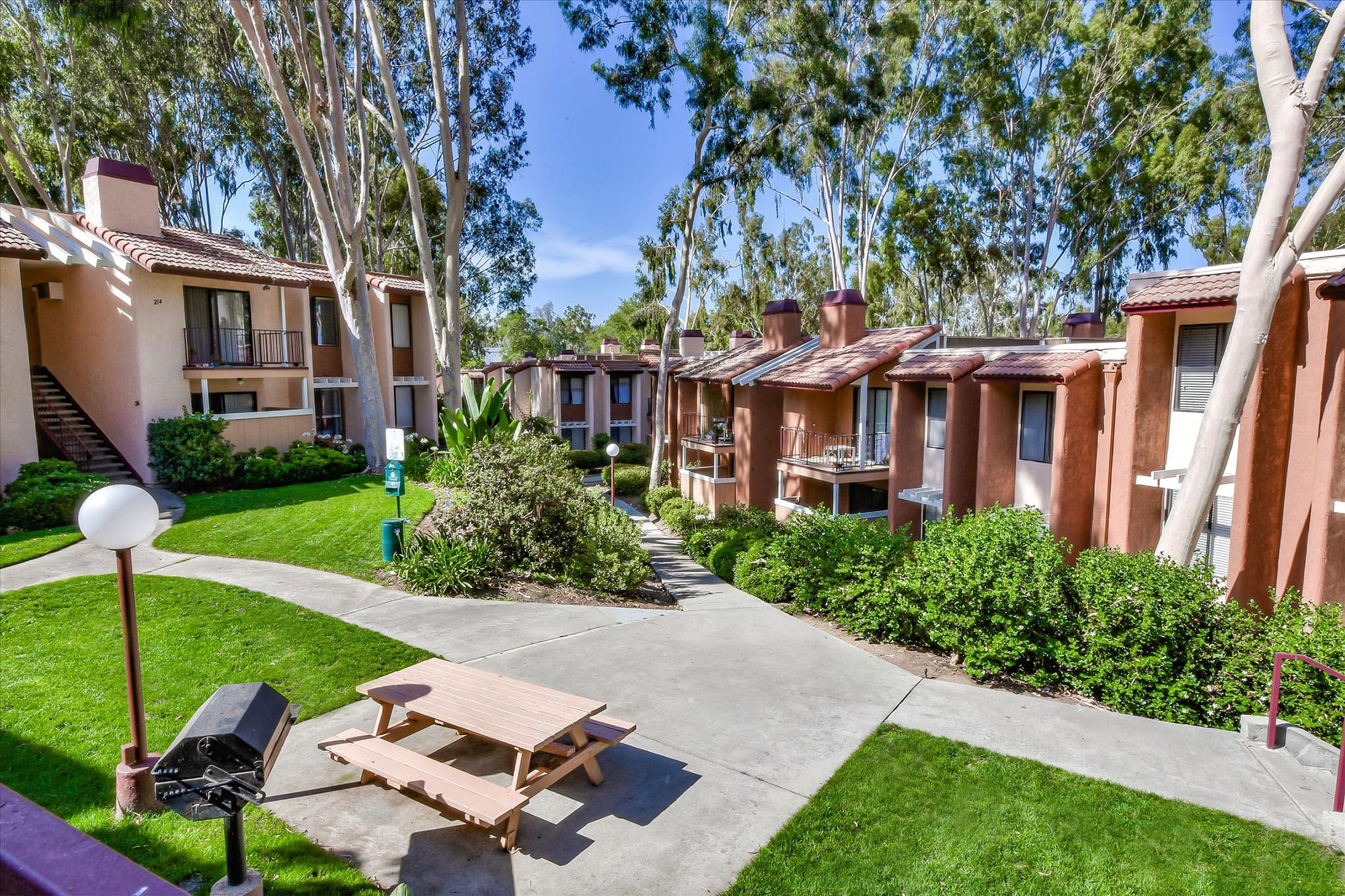 Exterior day shot of outside apartment complex, park seating, garden landscaping, and walking path