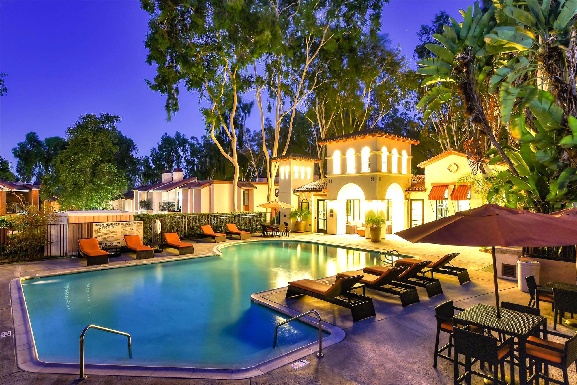 Exterior dusk shot of pool with tropical landscaping, shaded seating, and suntanning beds