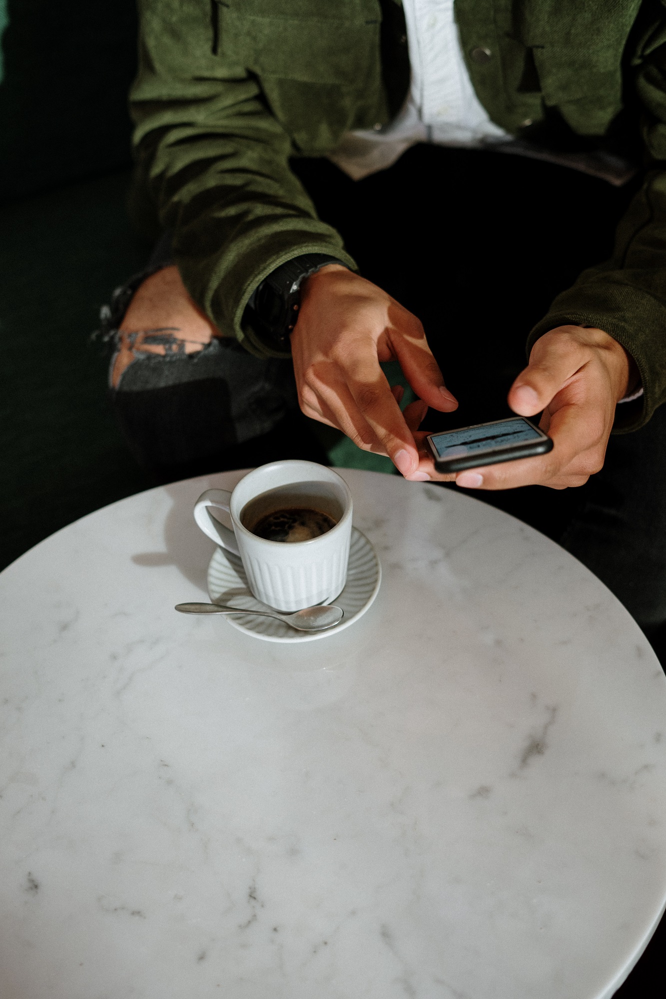 Young man sitting at coffee table on his phone with a cup of coffee and spoon