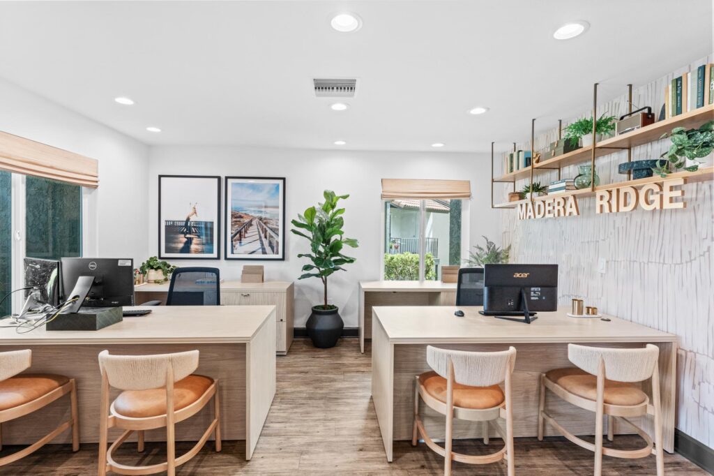Leasing office space with four desks, wooden chairs, and computers. Two framed photos on the wall, potted plants, wood flooring, and decorative shelving with "MADERA RIDGE" sign.