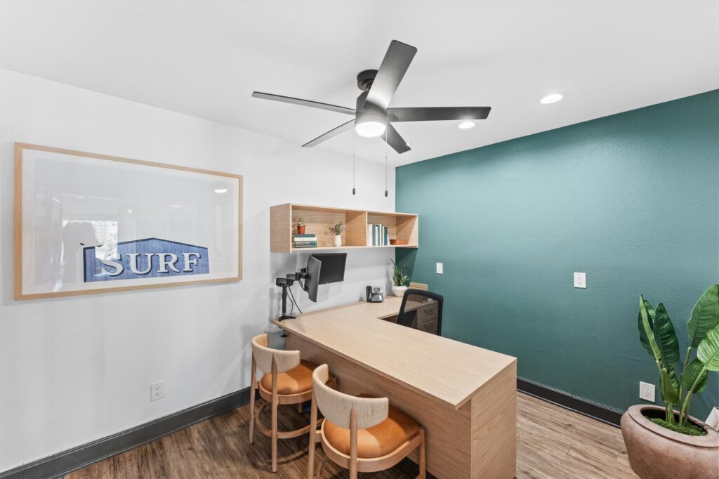 Leasing office with a wooden desk and two chairs, a teal accent wall, ceiling fan, and framed "SURF" art.