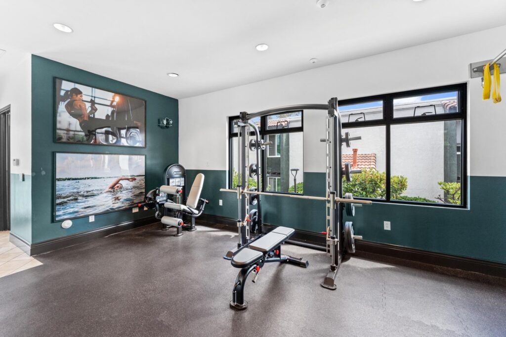 Gym with a weight bench and fitness equipment, bright natural light through large windows. Motivational fitness posters on the wall.