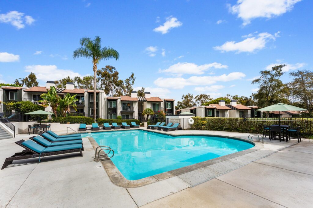 Sunny outdoor pool area with turquoise lounge chairs, surrounded by lush greenery and apartment buildings.