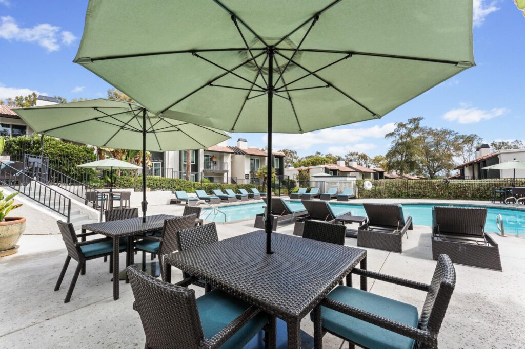 Sunny outdoor pool area with turquoise lounge chairs, surrounded by lush greenery and apartment buildings.