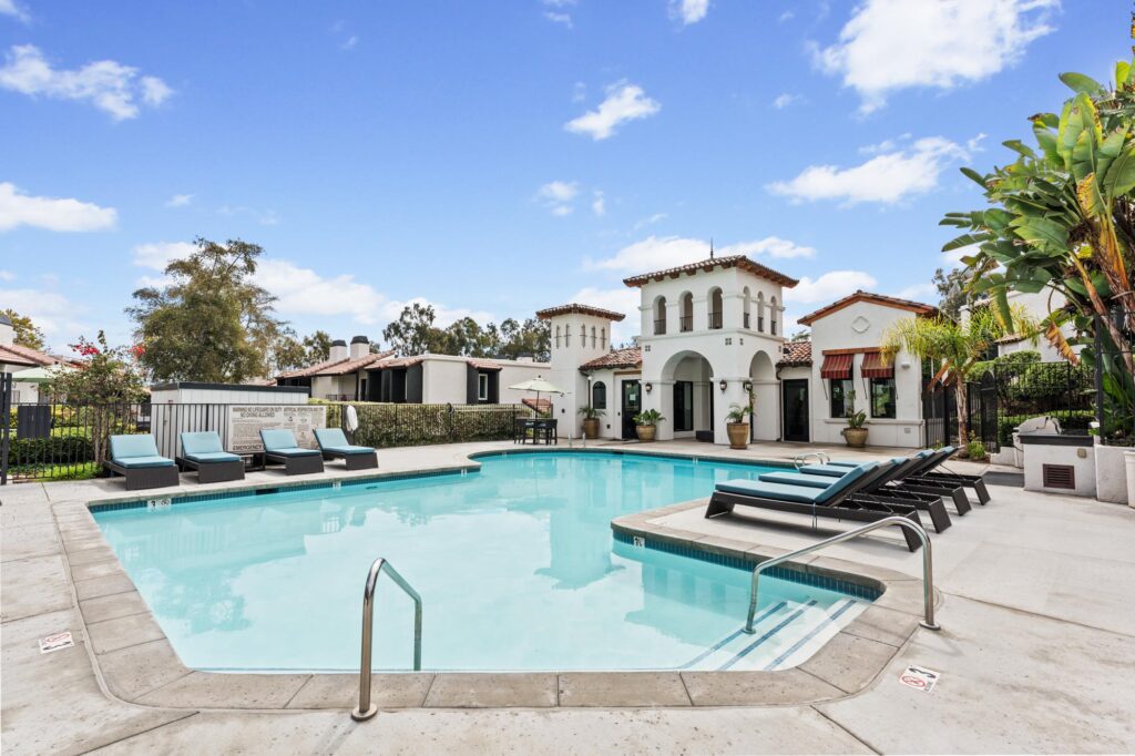 Outdoor pool surrounded by several turquoise lounge chairs on a stone patio. A Mediterranean-style clubhouse with arches and red-tiled roof is in the background.