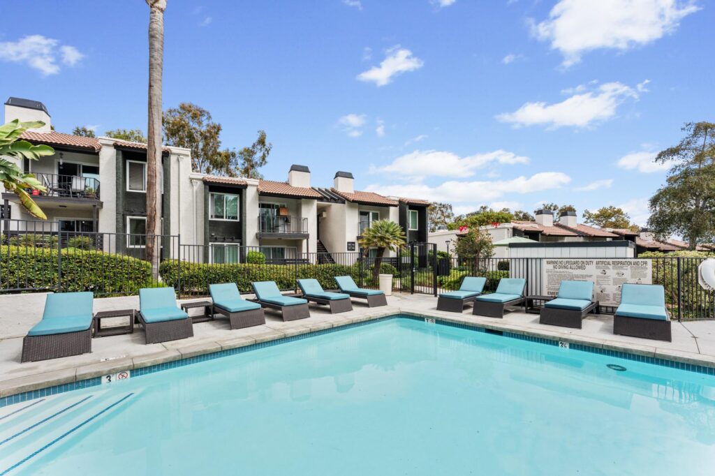Sunny outdoor pool area with turquoise lounge chairs, surrounded by lush greenery and apartment buildings.