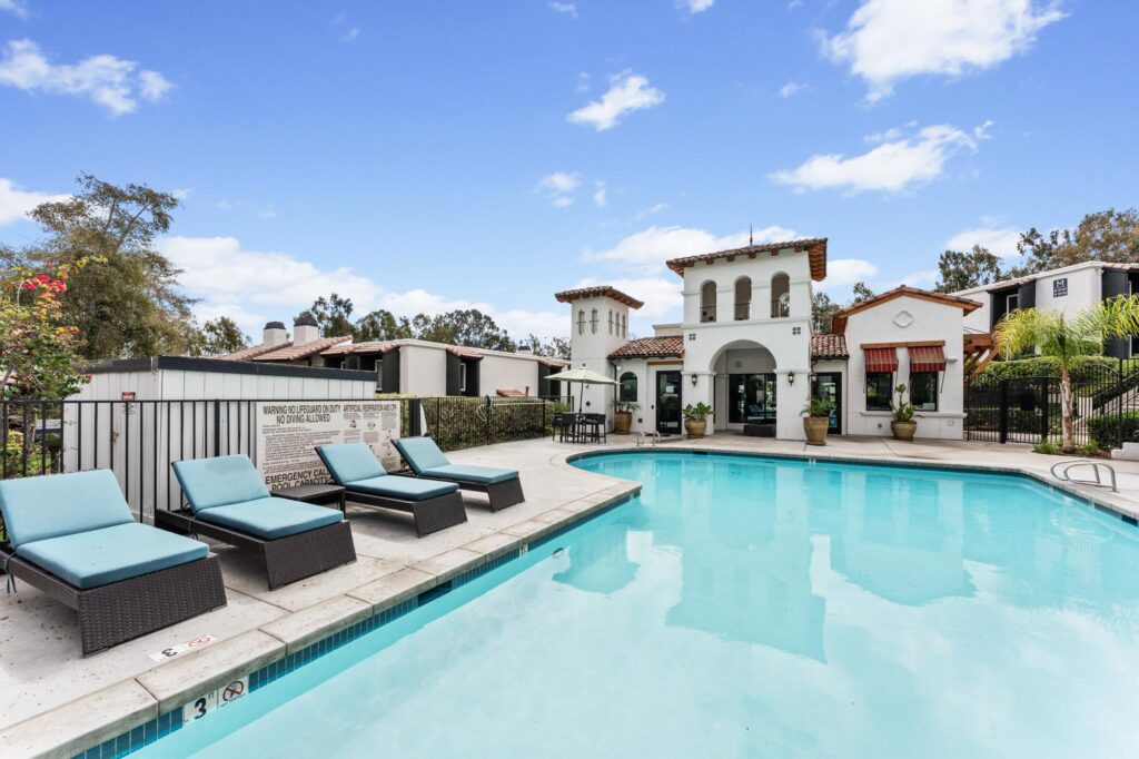 Outdoor pool surrounded by several turquoise lounge chairs on a stone patio. A Mediterranean-style clubhouse with arches and red-tiled roof is in the background.