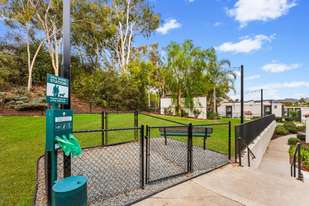 Fenced dog park with a waste station, including green bags, on a sunny day.